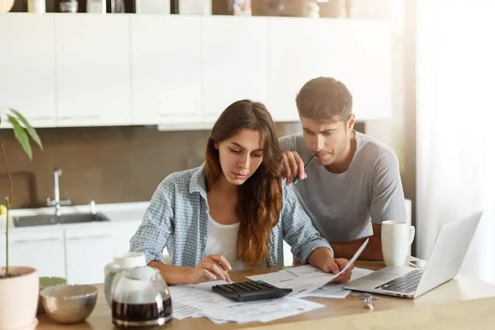 Busy  young woman calculating something on calculator, holding documents in hands while her husband leaning at her shoulder, keeping pen in mouth being preoccupied with financial report