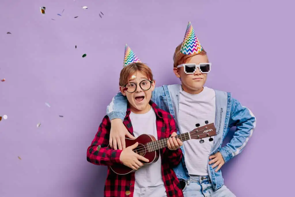 portrait of two friendly boys in casual wear stand posing at camera, birthday boy holding ukulele, guitar in hands. purple background