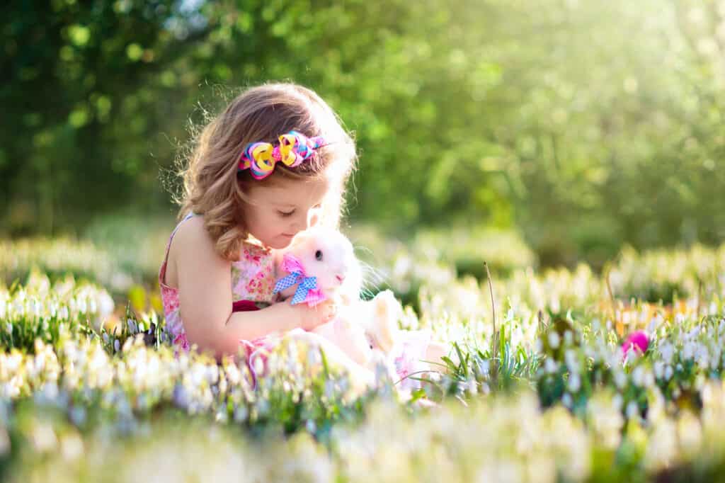 Adorable little girl, cute curly toddler in a colorful summer dress, playing with a real rabbit, having fun with her pet bunny in a beautiful garden with first spring snowdrop flowers