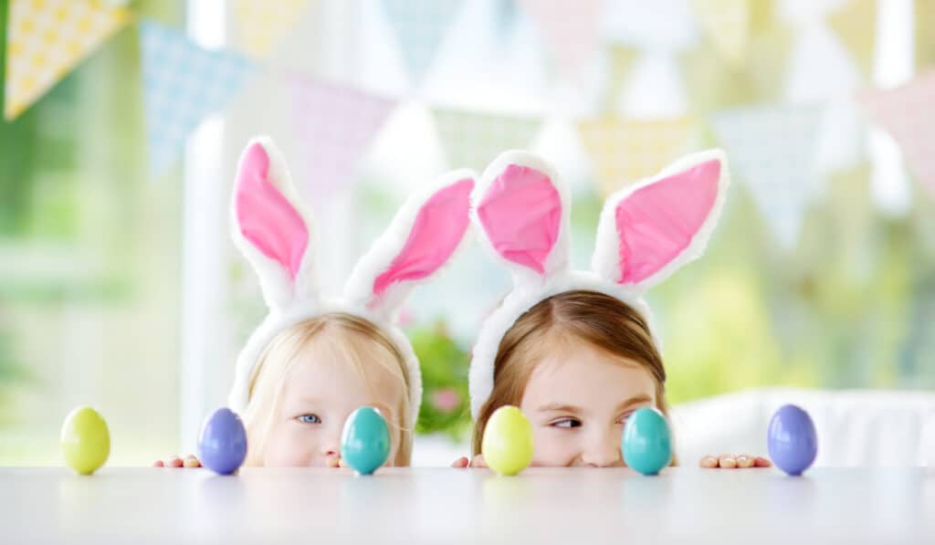 Two cute little sisters wearing bunny ears playing egg hunt on Easter. Adorable children celebrate Easter at home.